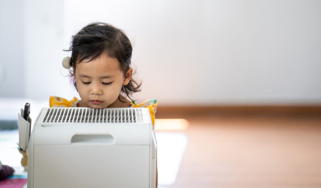 Shot of a little Asian girl standing near the portable air conditioner and curiously looking at it